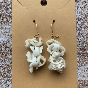 May include: A pair of off-white, abstract earrings with gold-colored ear wires. The earrings are made of layered, textured material, creating a unique, sculptural design. The earrings are displayed on a brown card.