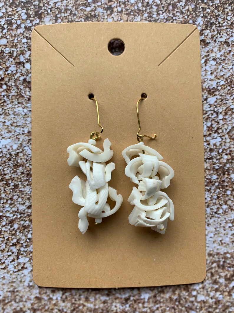 May include: A pair of off-white, abstract earrings with gold-colored ear wires. The earrings are made of layered, textured material, creating a unique, sculptural design. The earrings are displayed on a brown card.