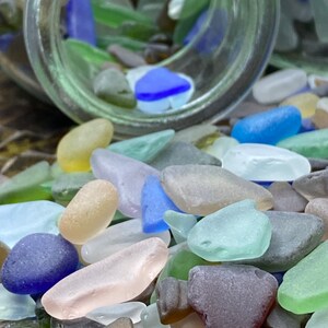 May include: A glass jar filled with colorful sea glass, spilling out onto a wooden surface. The sea glass is in various shapes and sizes, including blue, green, brown, and white.