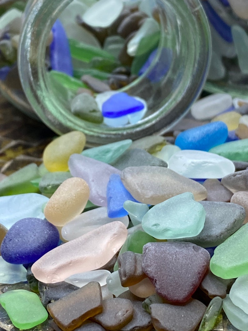 May include: A glass jar filled with colorful sea glass, spilling out onto a wooden surface. The sea glass is in various shapes and sizes, including blue, green, brown, and white.