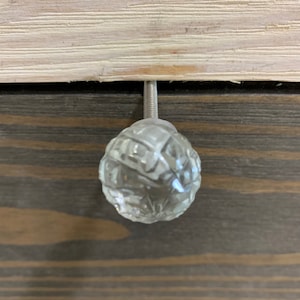 May include: A clear glass drawer knob with a faceted design. The knob is attached to a silver metal screw.