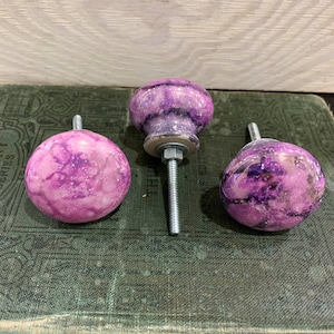 May include: Three decorative cabinet knobs with a marbled purple and white design. The knobs are round and have a metal screw for attachment. The knobs are displayed on a vintage green book.