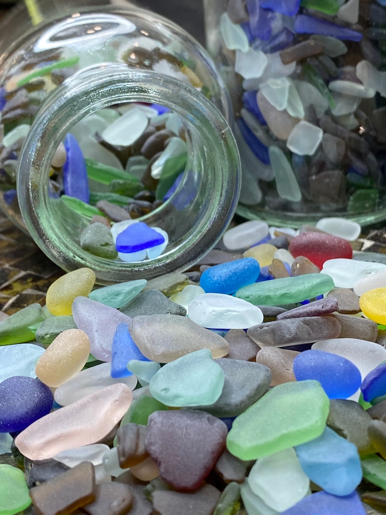 May include: A glass jar filled with colorful sea glass pieces. The sea glass is in various shapes and sizes, including small, medium, and large pieces. The sea glass is a mix of colors, including blue, green, brown, and white.