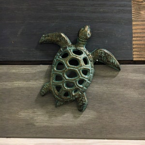 May include: A cast metal turtle wall decor with a distressed teal and gold finish. The turtle has a detailed shell design and outstretched flippers. The background is composed of horizontal wooden planks in shades of gray, black, and natural wood tones.