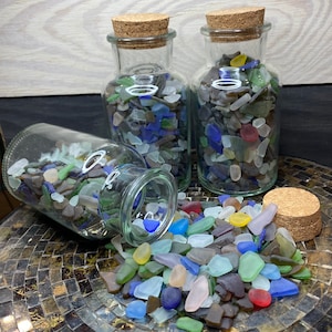 May include: Three glass jars filled with colorful sea glass. The jars have cork lids and are sitting on a tiled surface. Some of the sea glass has spilled out of one of the jars.