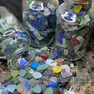 May include: Three glass jars filled with colorful sea glass. The jars are on a gold and brown patterned surface. The sea glass is a mix of blues, greens, yellows, and browns.