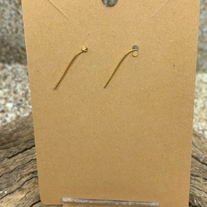 May include: A pair of gold-colored earring hooks displayed on a brown cardboard earring card. The card has a hole at the top for hanging and is held in place by a clear plastic stand. The background is a textured, neutral surface.