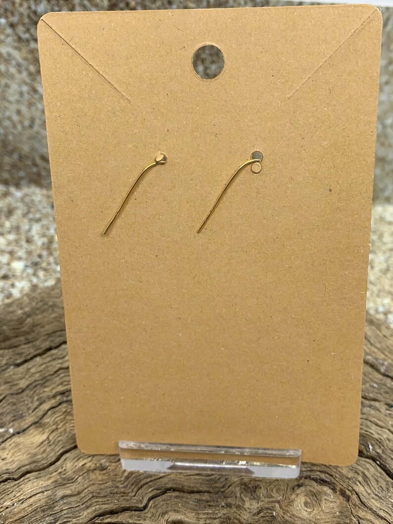May include: A pair of gold-colored earring hooks displayed on a brown cardboard earring card. The card has a hole at the top for hanging and is held in place by a clear plastic stand. The background is a textured, neutral surface.
