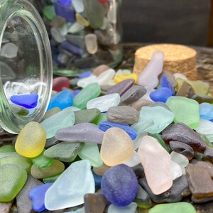 May include: A glass jar filled with colorful sea glass, spilling out onto a wooden surface. The sea glass is a mix of blues, greens, pinks, and browns.