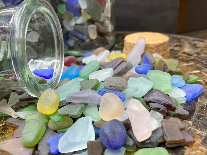May include: A glass jar filled with colorful sea glass, spilling out onto a wooden surface. The sea glass is a mix of blues, greens, pinks, and browns.