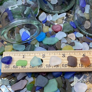 May include: A close-up of a collection of colorful sea glass pieces, some of which are arranged on a wooden ruler marked with inches. The sea glass is a variety of colors, including blue, green, brown, white, and red.