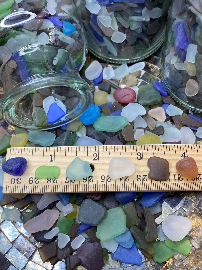 May include: A close-up of a collection of colorful sea glass pieces, some of which are arranged on a wooden ruler marked with inches. The sea glass is a variety of colors, including blue, green, brown, white, and red.