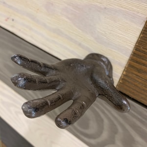 May include: A cast iron hand-shaped hook in a dark gray finish. The hook is mounted on a wooden surface.