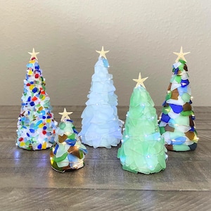 May include: Five decorative Christmas trees made of sea glass in various colors. Each tree is topped with a gold star and some are wrapped with fairy lights. The trees are arranged on a wooden surface.