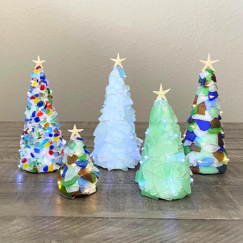 May include: Five decorative Christmas trees made of sea glass in various colors. Each tree is topped with a gold star and some are wrapped with fairy lights. The trees are arranged on a wooden surface.