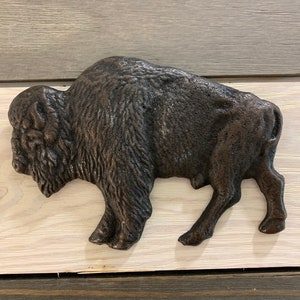 May include: A brown cast iron buffalo wall decor. The buffalo is standing with its head turned to the left. The buffalo is on a white wooden background.