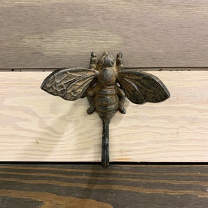 May include: A cast iron bee hook with a rustic brown finish. The hook is shaped like a bee with wings and a long tail that forms the hook.