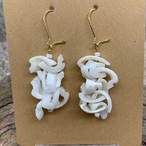 May include: A pair of white, abstract-shaped earrings with gold-colored ear wires. The earrings are made of layered, organic-looking pieces, creating a unique, sculptural design. They are displayed on a brown card.