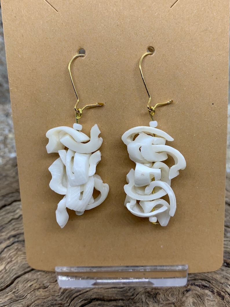 May include: A pair of white, abstract-shaped earrings with gold-colored ear wires. The earrings are made of layered, organic-looking pieces, creating a unique, sculptural design. They are displayed on a brown card.