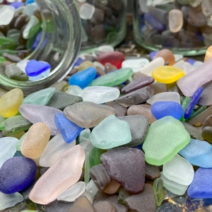 May include: A close-up of a variety of colorful sea glass pieces, including blue, green, yellow, and purple. The sea glass is scattered on a surface, with some pieces spilling out of a clear glass jar.