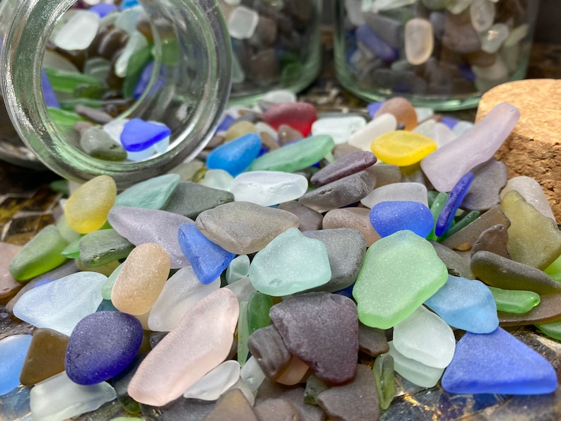 May include: A close-up of a variety of colorful sea glass pieces, including blue, green, yellow, and purple. The sea glass is scattered on a surface, with some pieces spilling out of a clear glass jar.