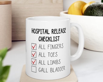 May include: White ceramic mug with the text "HOSPITAL RELEASE CHECKLIST". The checklist includes "ALL FINGERS", "ALL TOES", "ALL LIMBS", and "GALL BLADDER". The first three items are checked with red checkmarks.