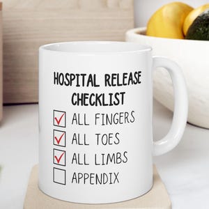 May include: White ceramic mug with the text "HOSPITAL RELEASE CHECKLIST" and check boxes for "ALL FINGERS", "ALL TOES", "ALL LIMBS", and "APPENDIX". The mug has a white handle and is on a beige coaster.