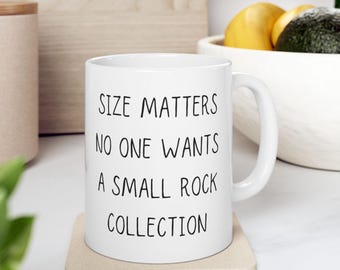May include: White ceramic mug with the text "SIZE MATTERS NO ONE WANTS A SMALL ROCK COLLECTION" in black. The mug is on a beige coaster, with a blurred background including a wooden cutting board and a bowl with fruit.