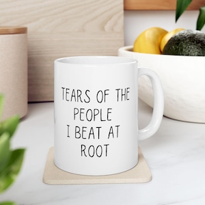 May include: White ceramic mug with the text "TEARS OF THE PEOPLE I BEAT AT ROOT" in black. The mug sits on a beige coaster. The background includes a wooden cutting board, a white bowl with fruit, and a wooden container.