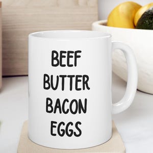 May include: White ceramic coffee mug with black text that reads "BEEF BUTTER BACON EGGS".