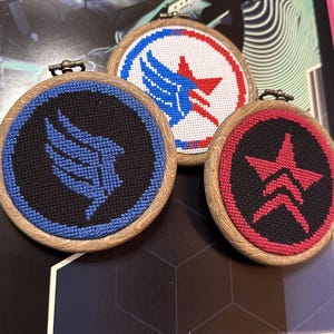 May include: Three circular wooden embroidery hoops containing cross-stitch designs. One features a blue wing on a black background, another a red star on a black background, and the third a blue and red wing design on a white background.