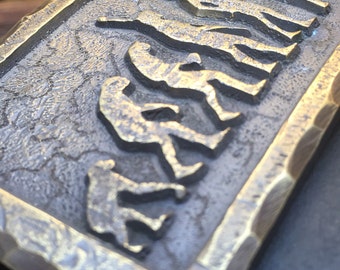 Puede incluir: Una hebilla de cinturón de metal dorado con un diseño en relieve de una serie de figuras en línea, que representan la evolución de los humanos.