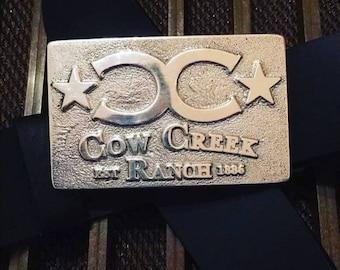 Puede incluir: Hebilla de cinturón de plata con un logotipo "CC" estilizado y dos estrellas. La hebilla también tiene el texto "Cow Creek Ranch Est 1886" grabado.