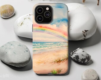May include: A phone case featuring a watercolor painting of a beach scene with a rainbow arching over the ocean. The case has a sandy beach, blue water, and a colorful sky. The case is surrounded by white stones and dried plants.