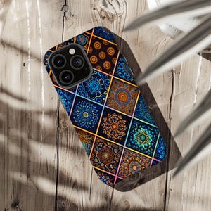 May include: A smartphone case with a vibrant geometric design. The case displays a pattern of squares with detailed, mandala-style motifs in blue, orange, and gold. The phone case is on a wooden surface, showing the camera array.