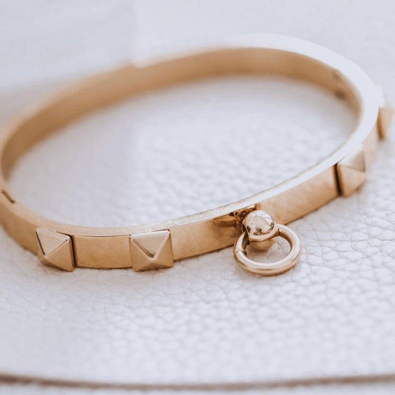 Puede incluir: Un brazalete dorado con un peque&ntilde;o charm redondo adjunto. El brazalete est&aacute; decorado con peque&ntilde;os tachuelas triangulares.