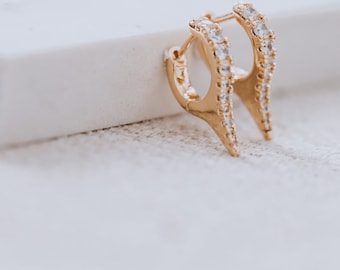 May include: Gold hoop earrings with a pointed design, featuring a row of clear cubic zirconia stones.