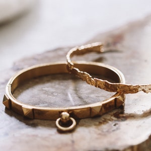 Puede incluir: Dos pulseras doradas sobre una superficie texturizada de color marr&oacute;n claro. Una pulsera tiene un acabado martillado y la otra tiene una fila de peque&ntilde;as p&uacute;as.