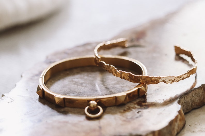 Puede incluir: Dos pulseras doradas sobre una superficie texturizada de color marr&oacute;n claro. Una pulsera tiene un acabado martillado y la otra tiene una fila de peque&ntilde;as p&uacute;as.