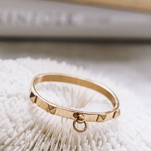 Puede incluir: Un brazalete dorado con un peque&ntilde;o charm redondo colgando. El brazalete est&aacute; decorado con peque&ntilde;os tachuelas triangulares.