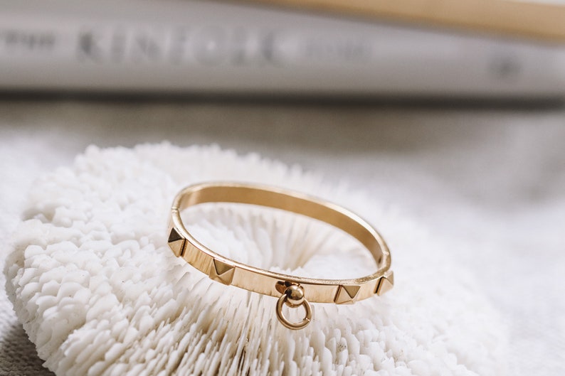 Puede incluir: Un brazalete dorado con un peque&ntilde;o charm redondo colgando. El brazalete est&aacute; decorado con peque&ntilde;os tachuelas triangulares.