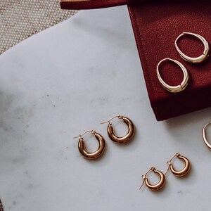 May include: A set of four gold hoop earrings of varying sizes. The earrings are displayed on a white marble surface and in a red box.