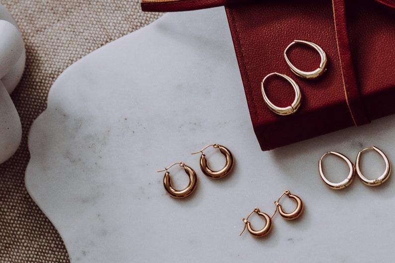 May include: A set of four gold hoop earrings of varying sizes. The earrings are displayed on a white marble surface and in a red box.