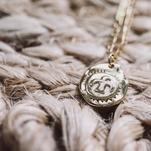 May include: A gold necklace with a round pendant featuring a stylized bird design. The pendant has a textured surface and a raised border.