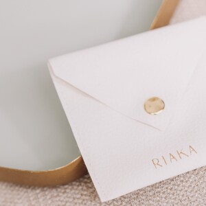 Pode incluir: Um envelope branco com fecho de bot&atilde;o dourado e a palavra "RIAKA" impressa em letras douradas.