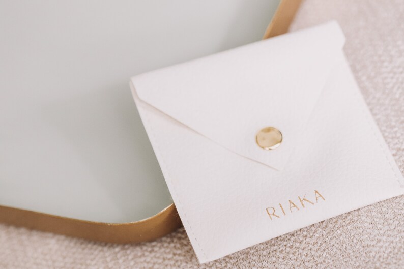 Pode incluir: Um envelope branco com fecho de bot&atilde;o dourado e a palavra "RIAKA" impressa em letras douradas.