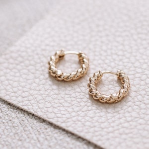 Peut inclure: Une paire de boucles d'oreilles créoles en or avec un design torsadé en forme de corde. Les boucles d'oreilles sont sur une surface texturée beige clair.