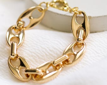 May include: A gold-colored chain bracelet with oval links and a lobster clasp. The bracelet is made of shiny metal and is displayed on a white surface. The links are connected by a small bar.
