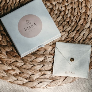 Pode incluir: Uma caixa branca com um logotipo circular rosa que diz "RIAKA Jewelry". A caixa est&aacute; sentada em um tapete de palha tran&ccedil;ado. H&aacute; um envelope branco com a palavra "RIAKA" impressa nele.