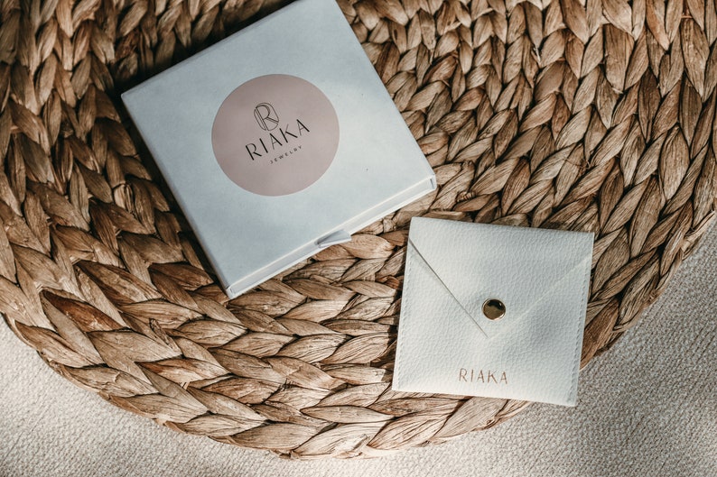 Pode incluir: Uma caixa branca com um logotipo circular rosa que diz "RIAKA Jewelry". A caixa est&aacute; sentada em um tapete de palha tran&ccedil;ado. H&aacute; um envelope branco com a palavra "RIAKA" impressa nele.